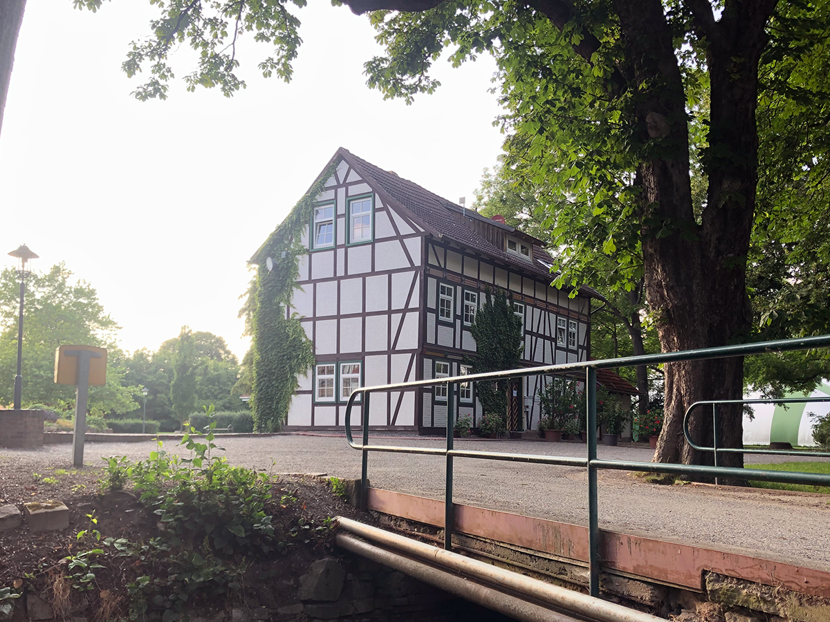 Ein Schotterweg führt über eine kleine Brücke zu einem von Bäumen umgebenen Fachwerkhaus.