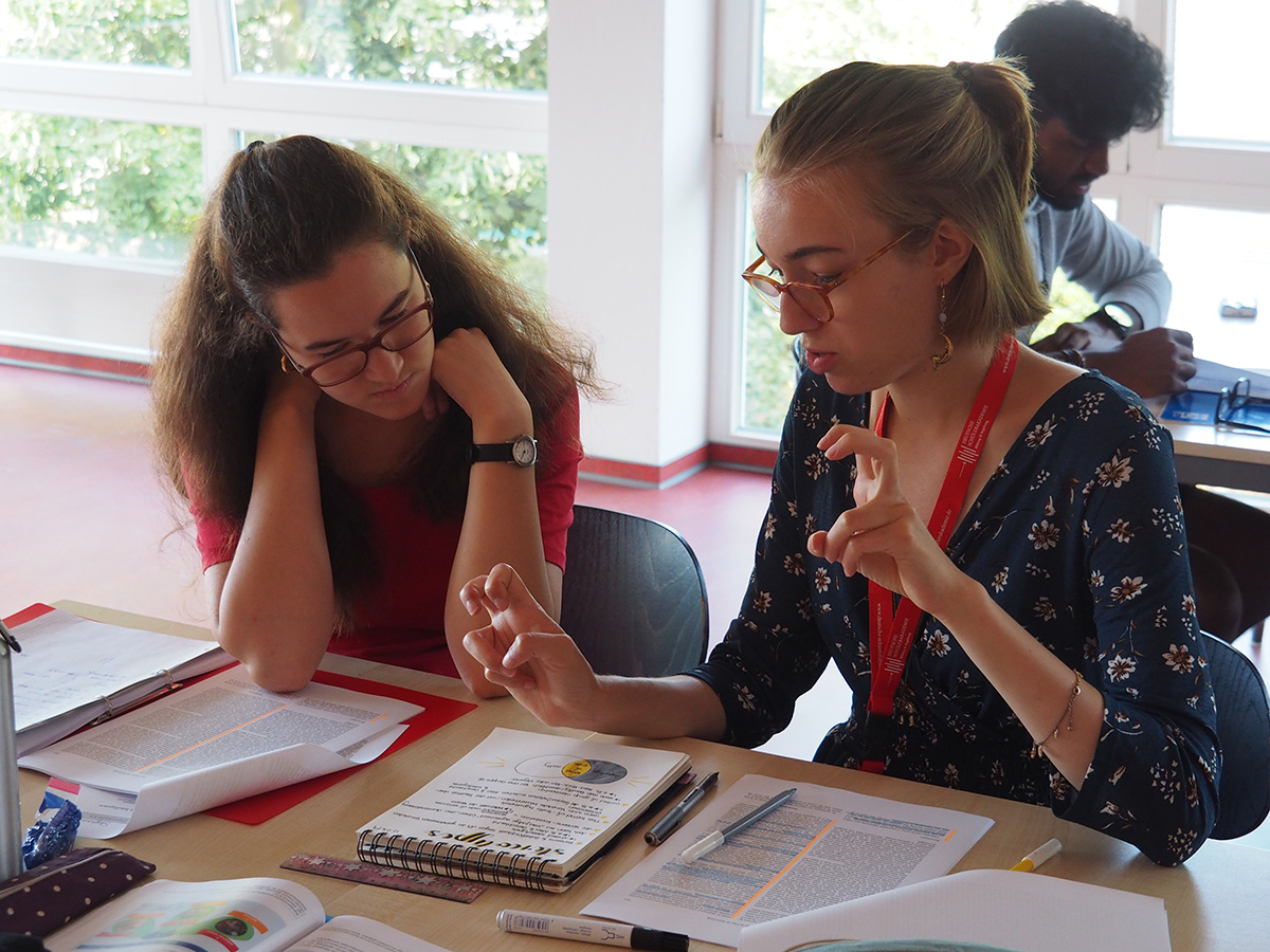 Zwei junge Frauen diskutieren über Unterlagen, die vor ihnen auf einem Schreibtisch liegen.