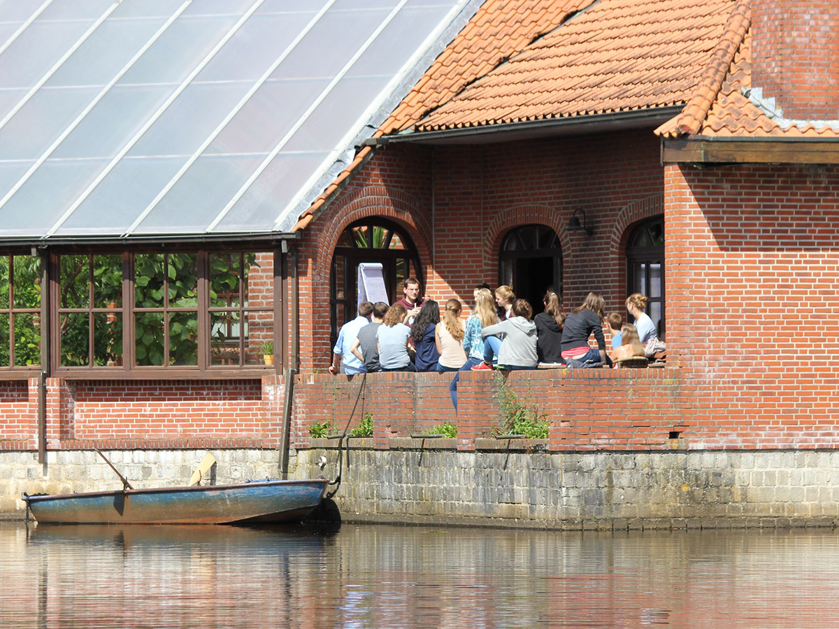 Von einem See aus gesehen sitzt eine Gruppe Jugendlicher auf einer halb überdachten Terasse bei einem Haus und schaut auf eine Flipchart. Am Haus liegt ein Boot.